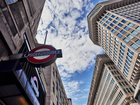 An artistic shot looking upwards, with the St James’ Park Underground sign on the left and the Ministry of Justice building on the right. The sky is blue with white clouds.