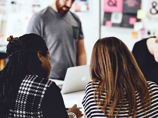 Three designers in the Design102 office. The backs of two women sitting and talking are in the foreground, and in the background a man stands, looking down at his MacBook screen.
