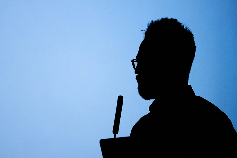 A dark silhouette of a man speaking into a microphone, against a light blue background.