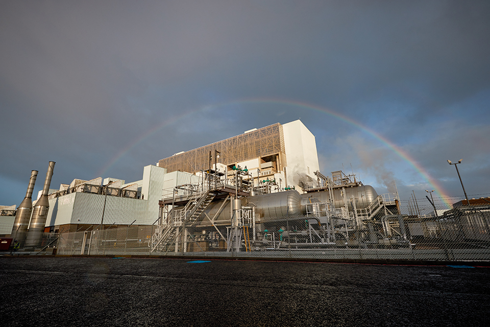 View of the power station from a road. A rainbow frames the building and the grey, stormy clouds are tinged with pink.