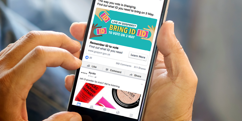 Somebody is holding a smart phone showing their Facebook feed. The phone says: Live in Gosport? Bring ID to vote on 3rd May.